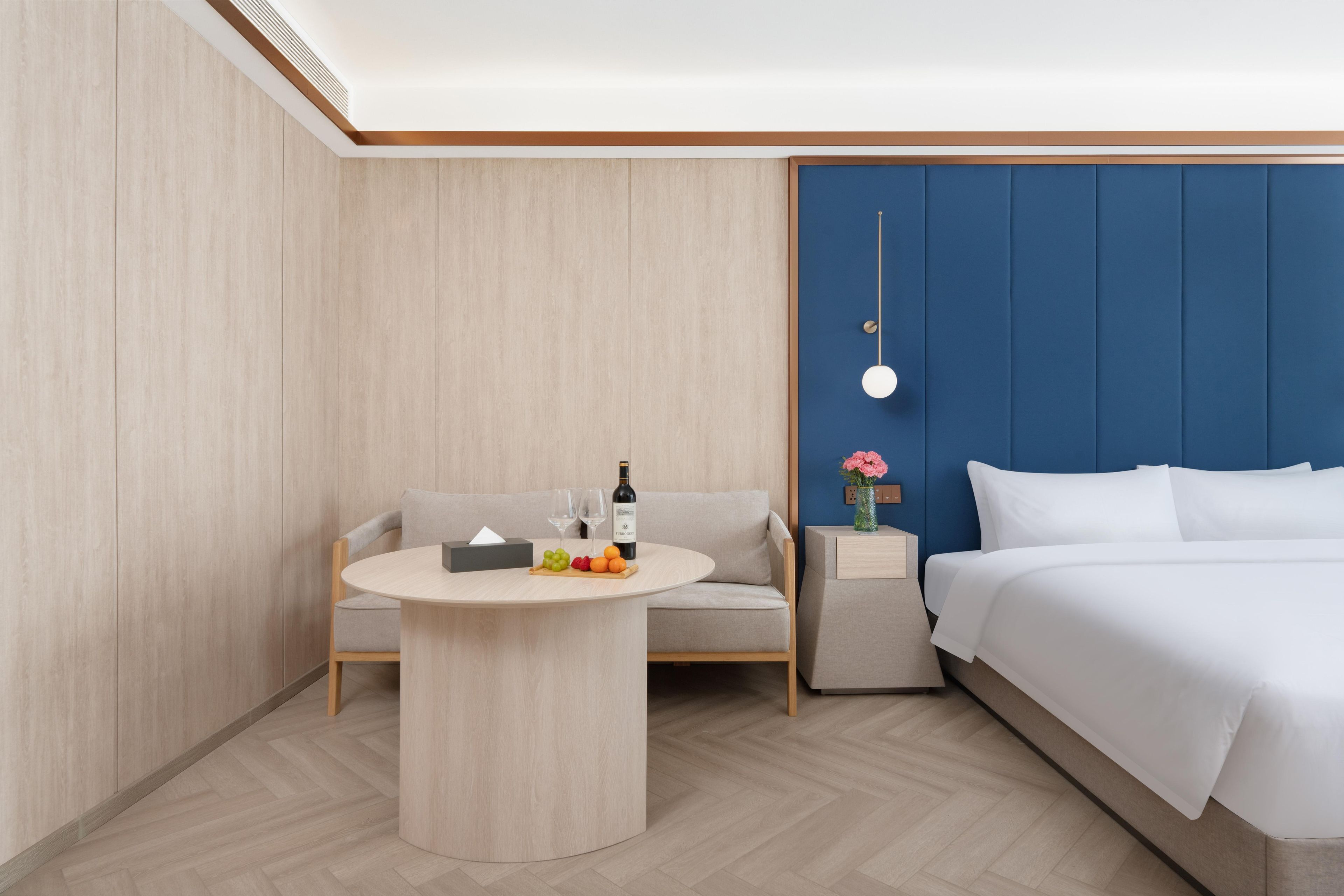 Habitación de hotel con una cama, una mesa redonda con frutas y una botella, un sofá y una mesita de noche con un florero.