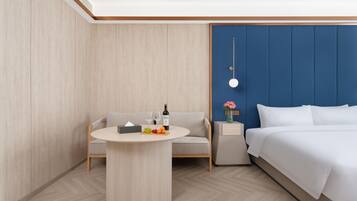 Habitación de hotel con una cama, una mesa redonda con frutas y una botella, un sofá y una mesita de noche con un florero.