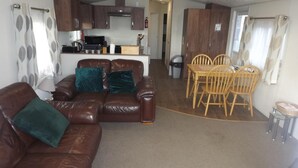 Un espacio habitable compacto con un sofá de cuero marrón, cojines color turquesa, una mesa de comedor de madera con cuatro sillas y una kitchenette al fondo.