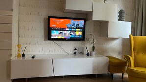 A modern living room with a flat-screen TV mounted on the wall, a white entertainment unit, a yellow armchair, and a small table with decorative items. - 1-Bed Apt, Sleeps 4, Central To Shops & Transport (London)
