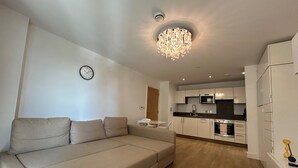 A modern living room with a beige sofa, a chandelier, a clock, and a kitchen in the background. - 1-Bed Apt, Sleeps 4, Central To Shops & Transport (London)
