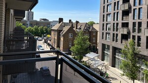 A multi-story building with a mix of residential and commercial spaces, featuring balconies, large windows, and a pedestrian street with cars and greenery. - 1-Bed Apt, Sleeps 4, Central To Shops & Transport (London)