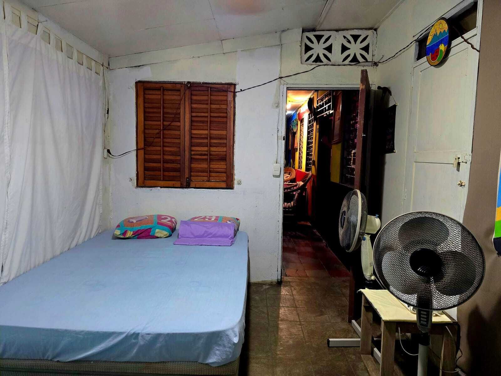 A small, narrow room with a single bed, a fan, and a window with wooden shutters.