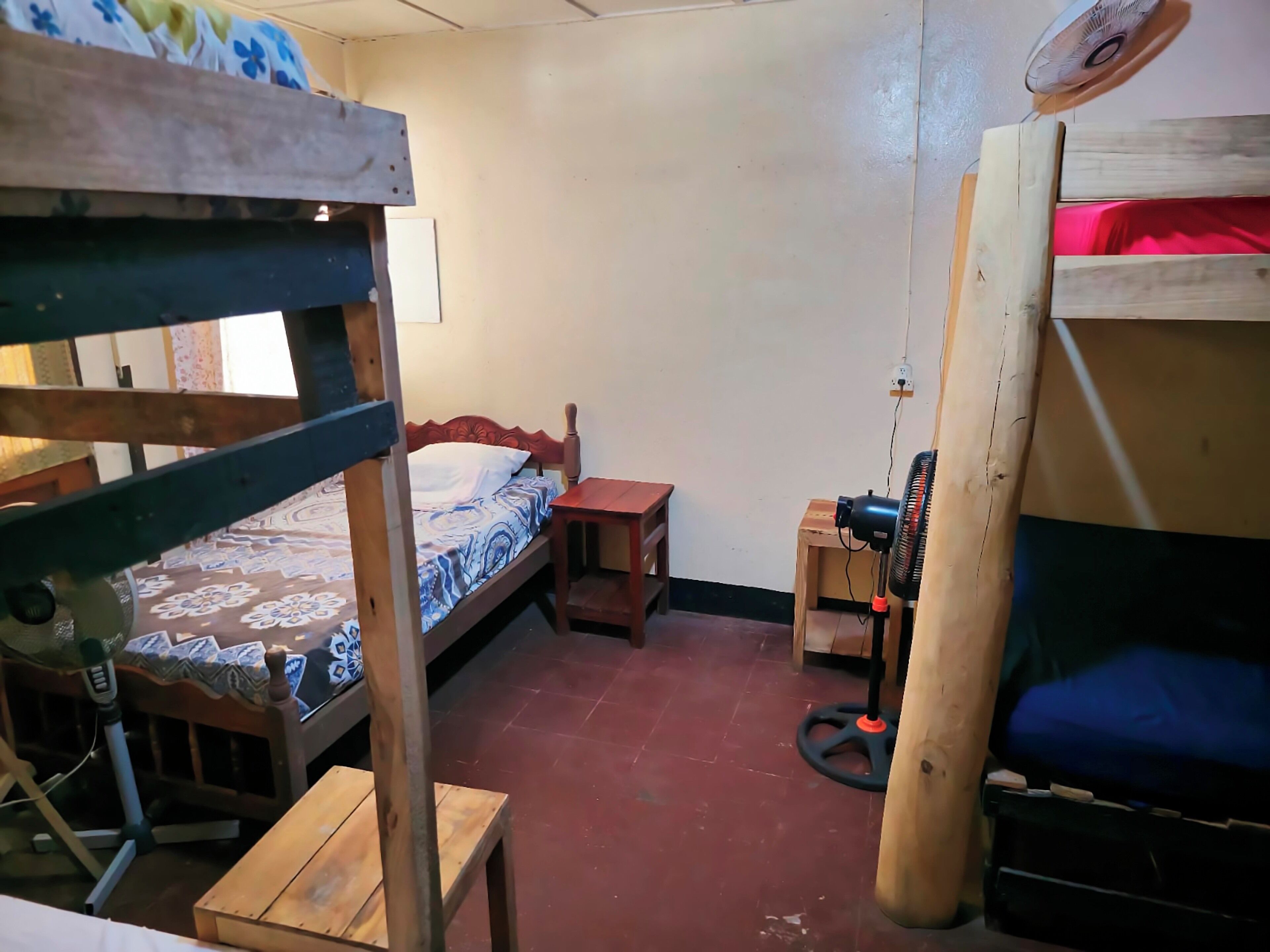 Un dormitorio con literas, una pequeña mesa de madera, un ventilador y una manta roja.
