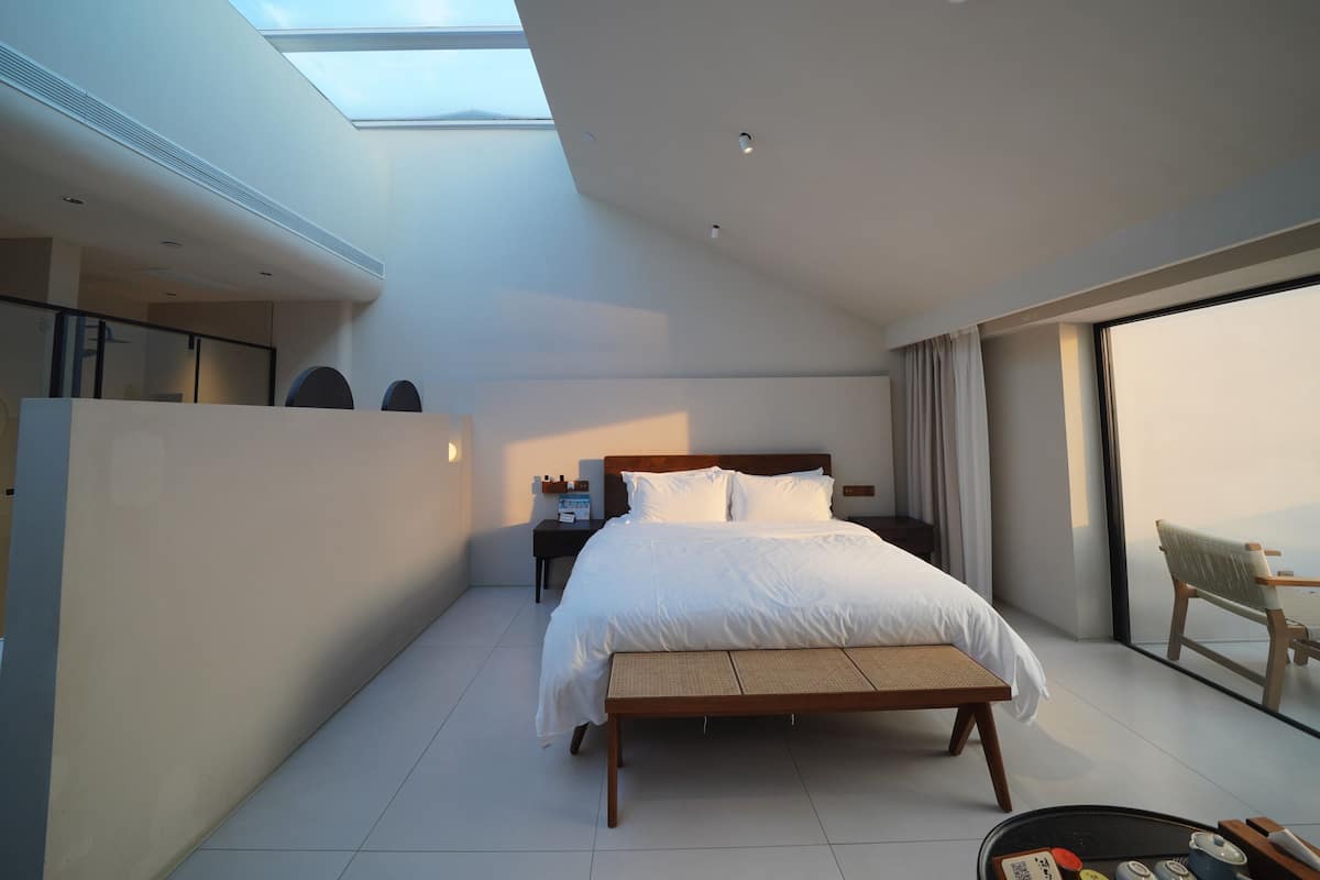 A modern hotel room with a skylight, a bed with white linens, a wooden bench, and a view of the sky.