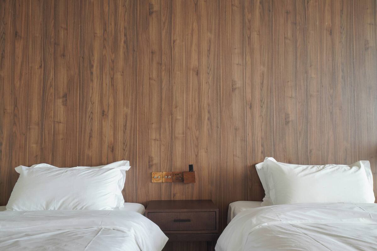 A hotel room with two single beds, wooden panel walls, a small wooden nightstand with drawers, and a wall-mounted light fixture.