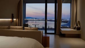 A modern hotel room with a large bed, a sofa, a small table, and a view of a lake at sunset. - Huyu Homestay (Suzhou)