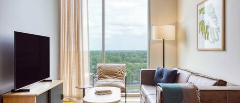 A modern hotel room with a large window, a flat-screen TV, a sofa, a chair, and a small table.