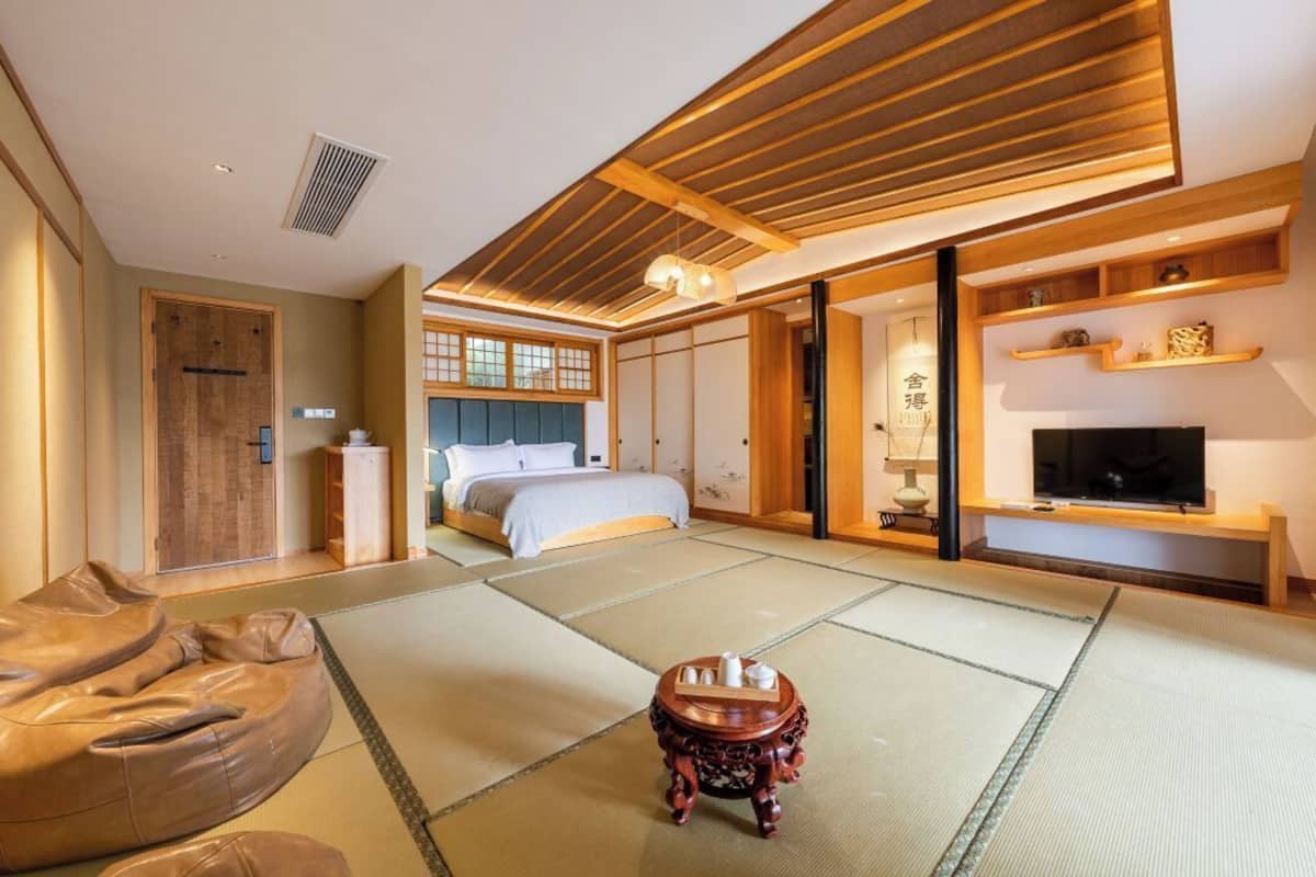 A traditional Japanese-style room with tatami flooring, a bed with a low headboard, a television on a wooden shelf, and a sliding door leading to another room.