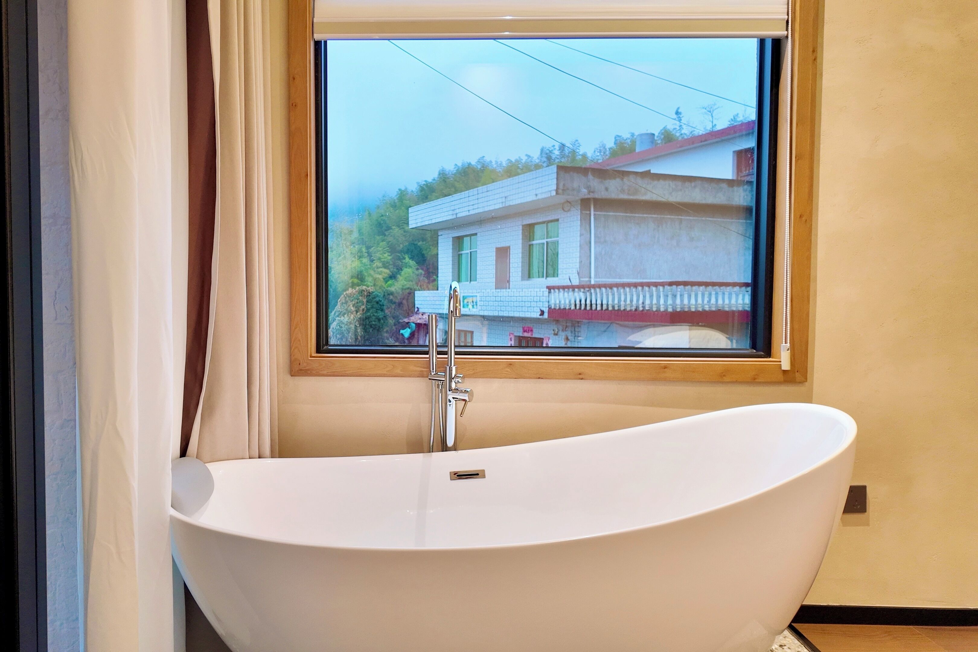A modern bathroom with a freestanding bathtub, a window with a view of a house, and a wooden-framed window.