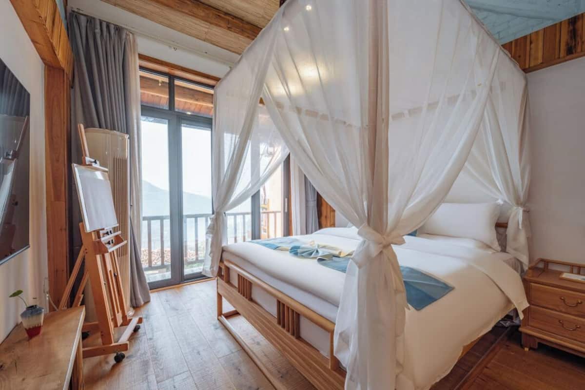 A bedroom with a four-poster bed, a wooden floor, a painting easel, and a large window.