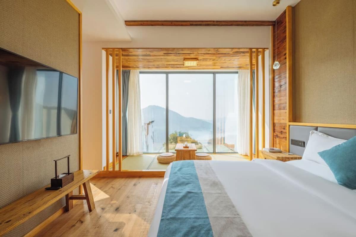 A modern hotel room with a large bed, a TV, a wooden bench, and a balcony with a view of the mountains.