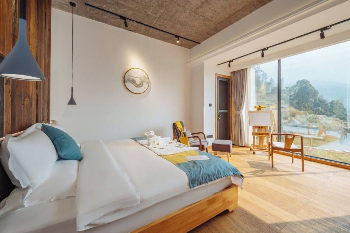 A modern bedroom with a large bed, a wooden headboard, a desk, and a chair. There is a large window with a view of the outdoors.