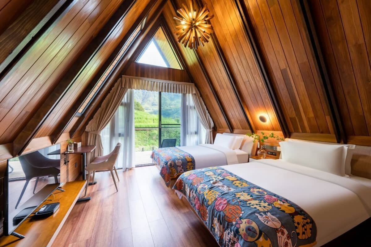 A cozy attic bedroom with a wooden sloped ceiling, a large bed with a colorful blanket, a desk with a chair, and a view of greenery outside.