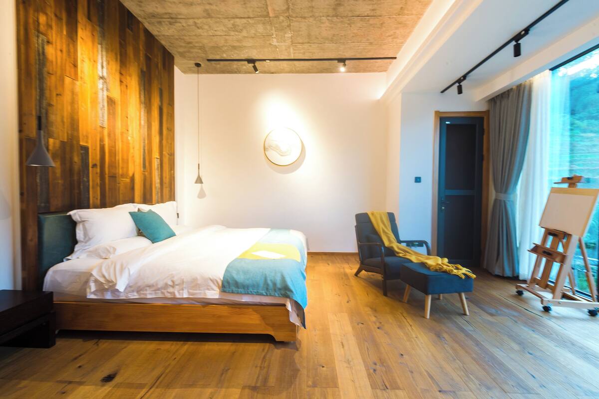 A modern bedroom with a wooden floor, a bed with white bedding, a blue chair, and an easel with a painting.