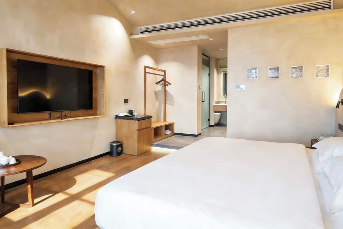 A hotel room with a large bed, a TV mounted on the wall, a small table with a cup, a hanging coat rack, and a bathroom visible through an open door.
