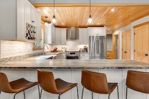 A modern kitchen with white cabinets, a marble countertop, stainless steel appliances, and wooden ceiling beams. - Slopeside Chalet | Hot Tub| Sauna| Game Room (Newry)