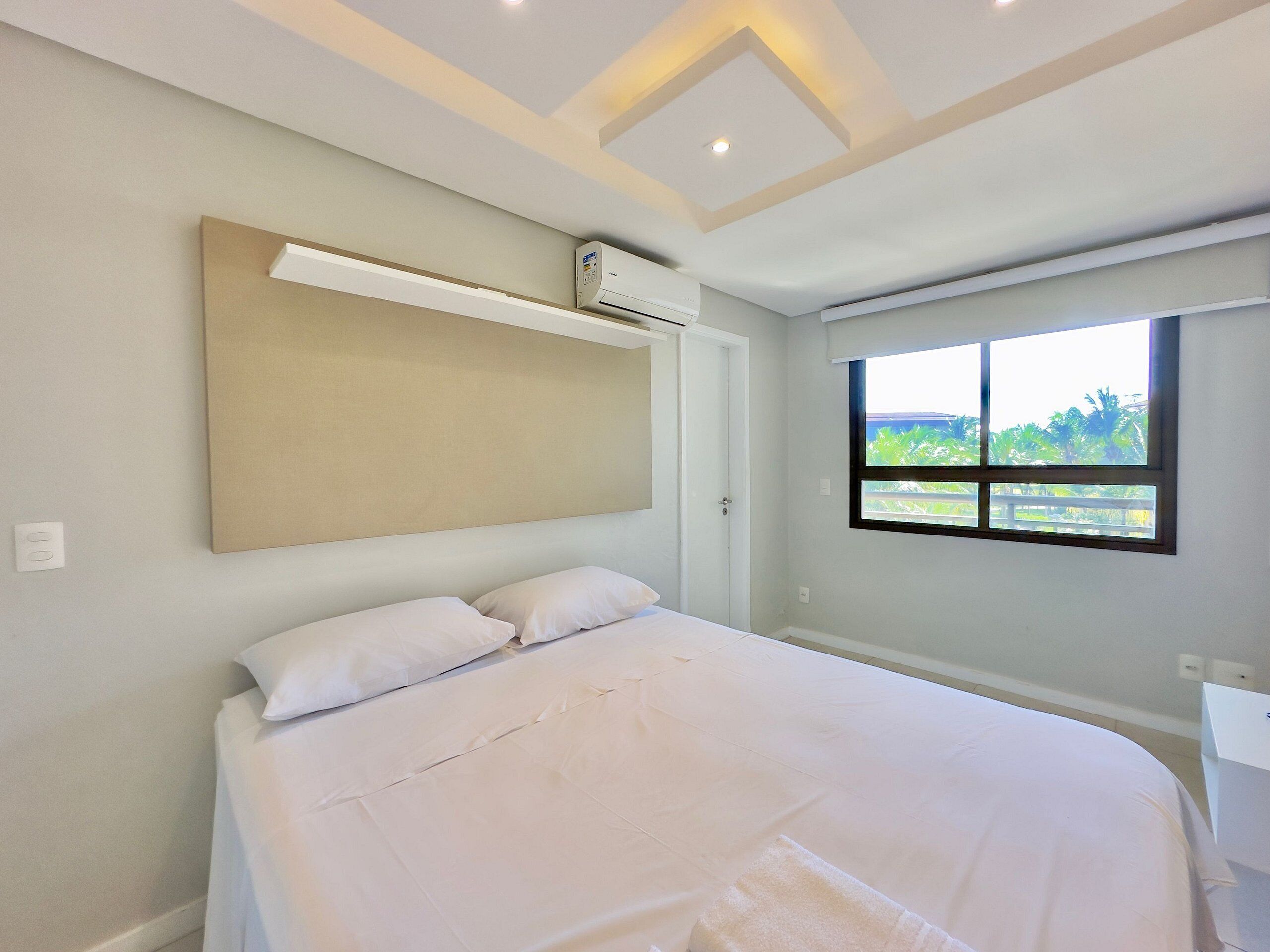 A bedroom with a bed, a window with a view, an air conditioning unit, and a light fixture on the ceiling.