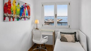 Une chambre avec un lit, un bureau et une chaise, une fenêtre donnant sur un port, et un tableau coloré représentant un oiseau accroché au mur.