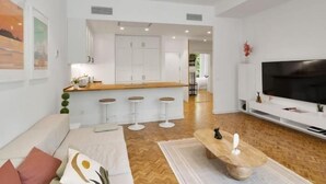 A modern living room with a sofa, a coffee table, and a TV. There is a kitchen area with a counter and stools.