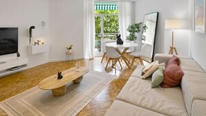 A modern living room with a flat-screen TV, a round dining table with chairs, a sofa with cushions, and a large window with a view of greenery.