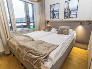 A double bed with a beige blanket, two pillows, and a headboard with a light fixture.