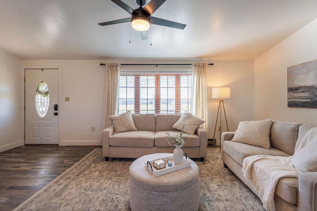 A living room with a beige sofa, a round ottoman, a ceiling fan, and a window with blinds.