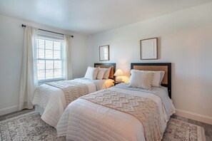 Une chambre avec deux lits, une fenêtre avec des rideaux et des œuvres d’art encadrées accrochées au mur.