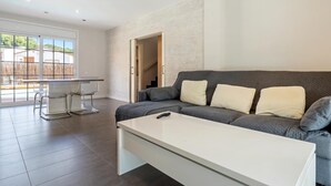 Un salon moderne avec un canapé gris, une table basse blanche et des chaises en verre transparent. Un escalier mène à un niveau supérieur, et une grande fenêtre offre une vue sur l’extérieur.