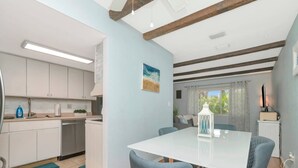 A modern kitchen with white cabinets, a dining table with chairs, and a view of the outdoors through a window. - The Largo Coastal Cottage (Largo)