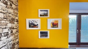 Une pièce avec un mur en pierre, un mur jaune et une fenêtre avec des stores. Quatre tableaux encadrés sont accrochés au mur jaune.