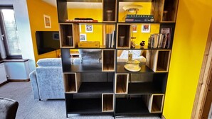 Un salon avec un canapé d’angle, une table basse et une bibliothèque adossée à un mur jaune.
