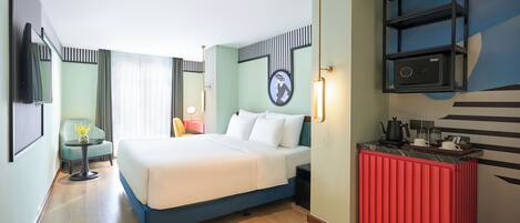 A hotel room with a bed, a small table, a chair, a red minibar, and a wall-mounted TV.