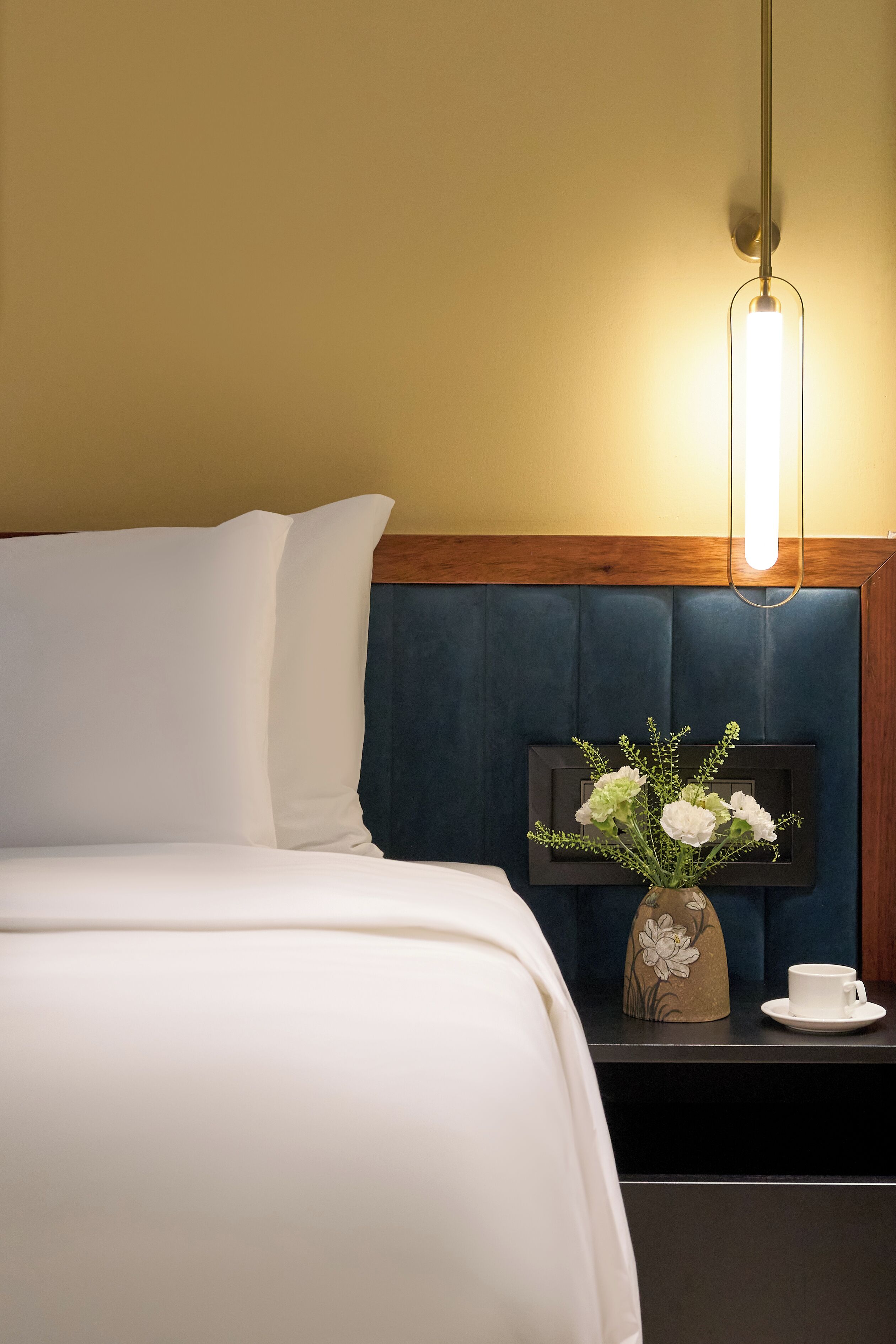 A hotel room with a bed, a nightstand, a vase with flowers, and a cup.