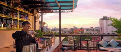 A bar area with a bartender preparing drinks, a bar counter, stools, and a view of the cityscape at sunset.