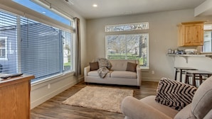 A spacious living room with a sofa, a coffee table, and a rug. There are large windows with blinds, a kitchen area with wooden cabinets, and a view of the outdoors. - Convenient 3BR with King Beds Near Hill AFB and Davis Conference Center (Layton)