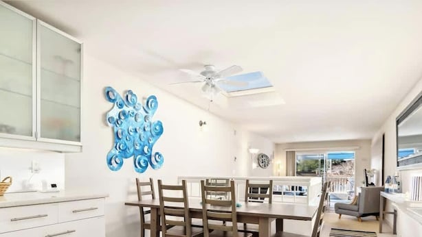 A modern kitchen with white cabinets, a dining area with wooden chairs, a ceiling fan, and a large window providing a view of the outdoors. - Capitola Sands (Capitola)