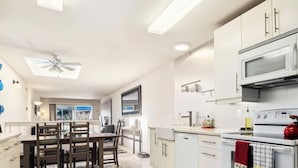 A modern kitchen with white cabinets, a microwave, an oven, and a red kettle. There is a dining table with chairs and a ceiling fan. - Capitola Sands (Capitola)