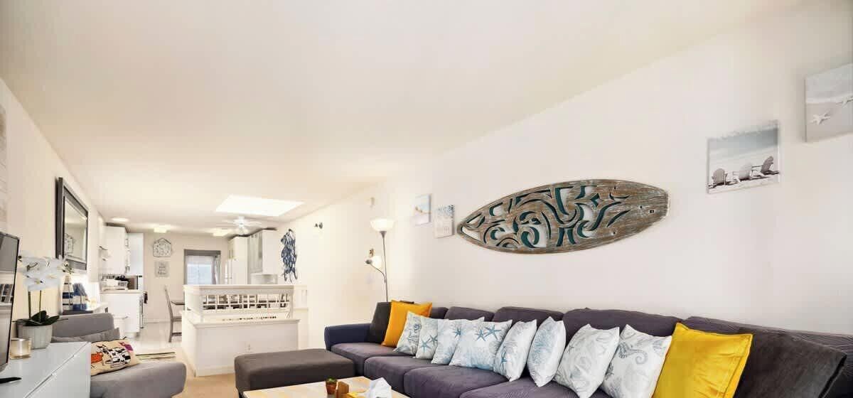 Ein modernes Wohnzimmer mit einem dunklen Sofa, mehreren Kissen, einem Couchtisch und einer Küche im Hintergrund.
