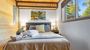 A bedroom with a wooden ceiling, a large bed with a grey headboard, two bedside tables with lamps, and a window with a view of trees. - Gooseberry Trailside Suites | Encampment River Suite (Two Harbors)