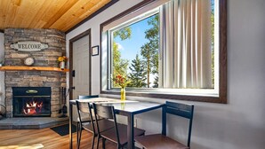 A cozy room with a stone fireplace, a dining table with chairs, and a large window with curtains. - Gooseberry Trailside Suites | Encampment River Suite (Two Harbors)