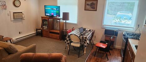 A living room with a brown couch, a dining table with chairs, a TV, and a kitchen area with a stove.