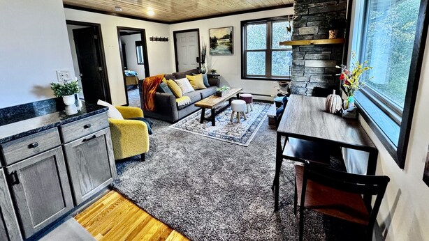 A modern kitchen with a stone accent wall, a dining area with a wooden table, and a cozy seating area with a sofa and chairs. - Gooseberry Trailside Suites | Lester River Suite (Two Harbors)