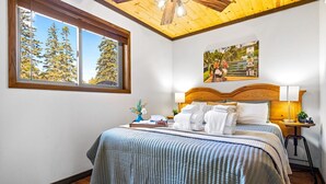 Una cama bien hecha con una colcha azul y blanca a cuadros, un cabecero de madera y dos lámparas de noche. Se ve una ventana con vistas a árboles y un cuadro en la pared.
