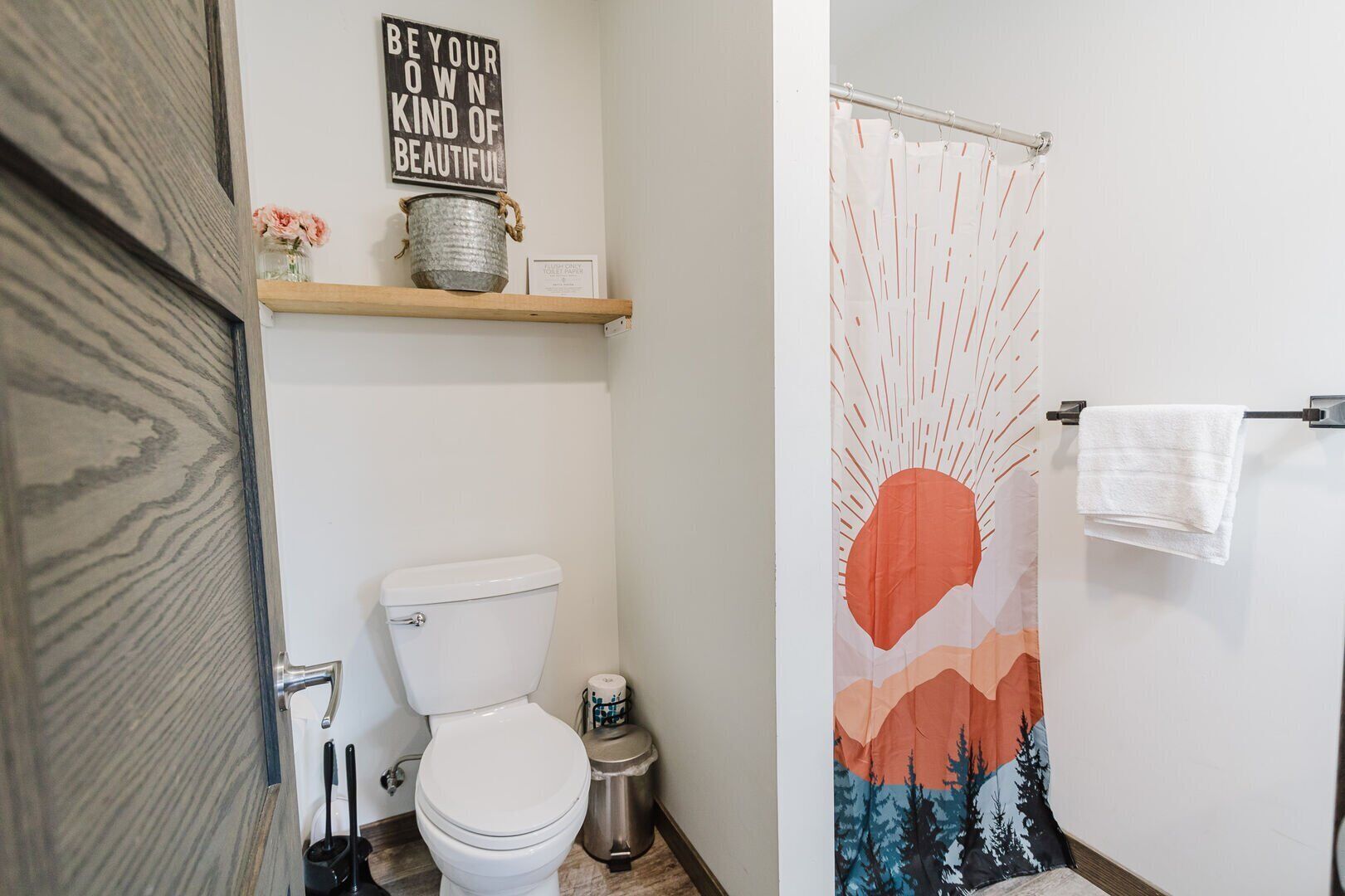 A bathroom with a toilet, a shelf with a sign, a trash can, and a shower curtain with a mountain design.