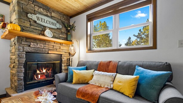 Ein gemütliches Wohnzimmer mit Kamin aus Stein, einem grauen Sofa mit bunten Kissen, einem Holztisch und einem Fenster mit Blick auf Bäume.