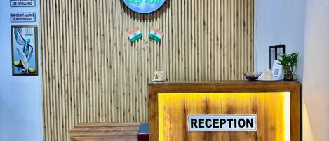 A reception desk with a wooden finish and a "RECEPTION" sign. Above, there is a circular sign with the hotel's logo and name.