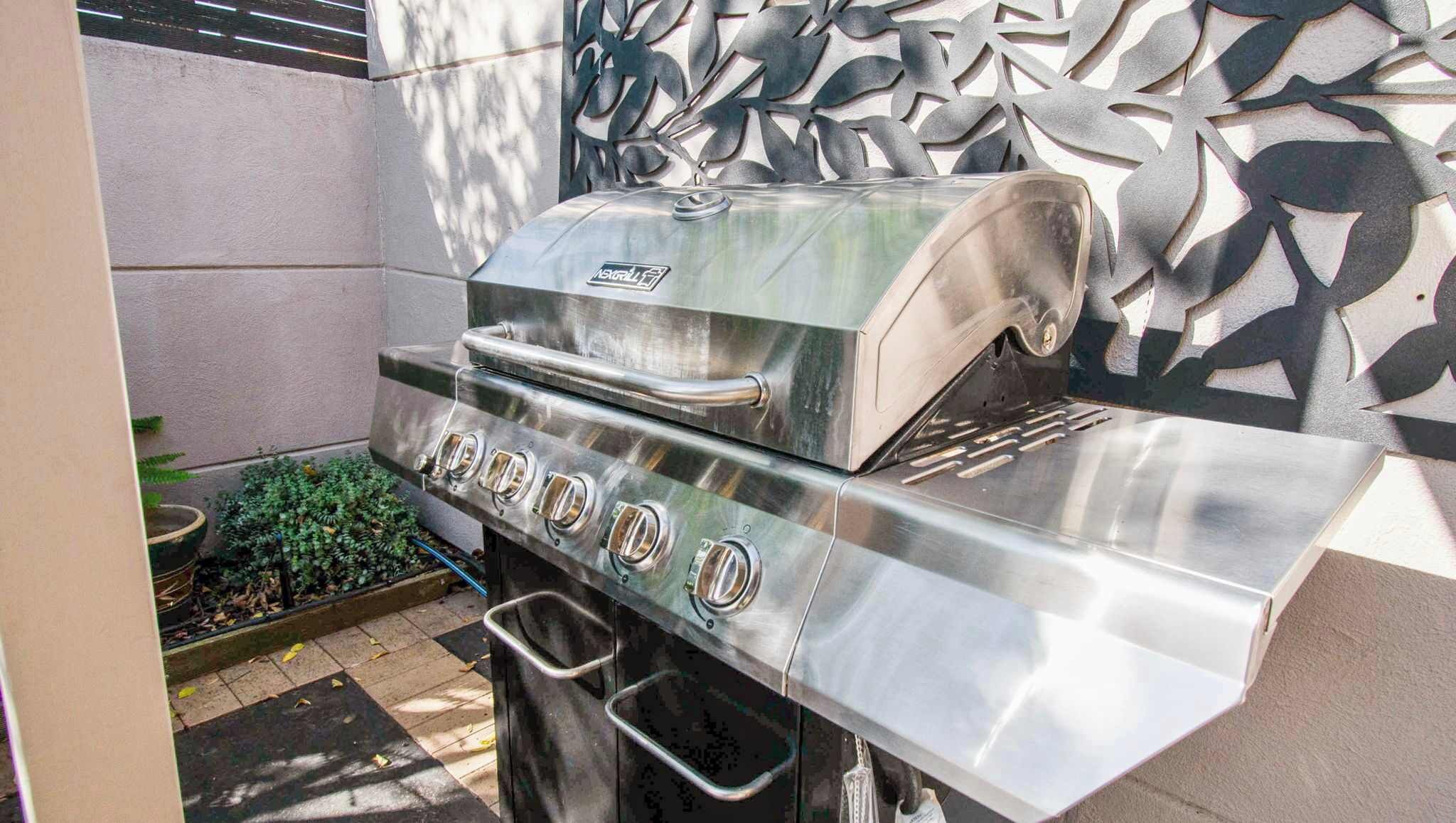 A stainless steel outdoor grill with a digital display, located in a patio area with a decorative wall and a small garden.