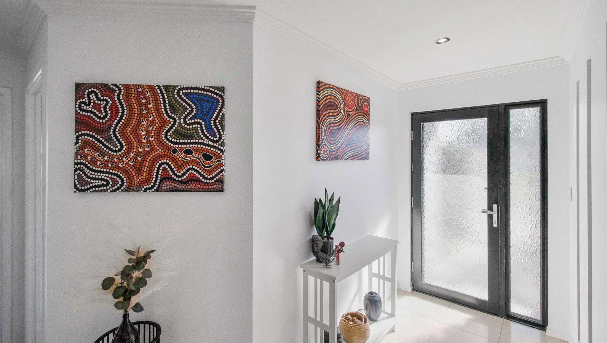 A modern interior with two wall-mounted artworks, a potted plant, and a glass door leading to a bright room.