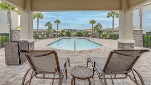 A covered pool area with lounge chairs and a table, surrounded by palm trees and a view of residential buildings. - Golfer’s Paradise! Extended-Stay Bradenton Condo (Bradenton)